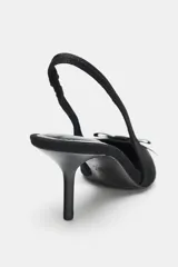 Zapato de tacón destalonado tipo slingback, confeccionado en tejido negro. Presenta punta fina, un pequeño lazo decorativo en la parte delantera y una tira elástica en el talón para mayor ajuste.