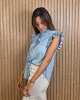 Musculosa de jean celeste sin mangas, con cuello alto fruncido, cierre frontal con botones y detalles de volados en las sisas y a lo largo de la botonera.