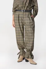 Pantalon de corte holgado con estampado a cuadros en tonos marrones y verdes. Presenta pinzas en la cintura, bolsillos laterales y un diseño distintivo en los tobillos con tiras ajustables para anudar.