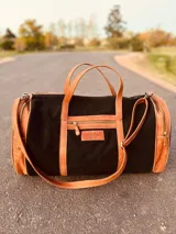 Bolso de viaje grande de cuero vacuno color negro con detalles en marrón. Tiene herrajes metálicos, gran superficie interna, asa de mano y correa para el hombro.