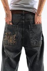 Pantalón de denim azul desgastado, corte mega baggy con piernas anchas y bolsillos traseros grandes. Presenta un bordado dorado en estilo gótico en el bolsillo trasero izquierdo.
