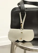 Estuche para lentes de ecocuero negro con textura granulada, diseño minimalista con cierre de broches metálicos dorados y correa de mano desmontable.