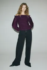 Blusa de color violeta con diseño de hombros descubiertos, cuerpo fruncido y mangas largas abullonadas con puños elásticos. Presenta detalles de volados en el escote, el bajo y los puños.