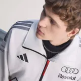 Campera adidas del equipo Audi Revolut F1 Team, de color blanco con cierre frontal negro y cuello alto. Presenta detalles de patrocinadores estampados en el pecho y las mangas, junto con las clásicas tiras de adidas en los hombros.
