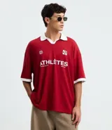 Remera de manga corta estilo polo, color rojo, con cuello y puños en contraste blanco. Presenta un corte oversized con hombros caídos y la palabra "ATHLÈTES" estampada en blanco en el frente. Incluye pequeños gráficos blancos a cada lado del pecho.