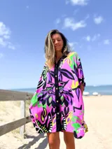 Camisero corto de corte holgado con estampado floral vibrante en tonos lila, fucsia, verde y negro. Presenta cierre frontal con botones, cuello camisero y mangas cortas.