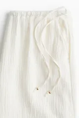 Pantalón de corte holgado color blanco, con piernas amplias y bolsillos al bies. Modelo de tiro bajo con elástico revestido y cordones para ajustar enlazados a los lados, decorados con cuentas en los extremos.
