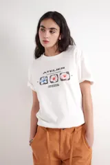 Remera de corte oversize, color blanco, con cuello redondo y mangas cortas. Presenta una estampa frontal con el texto 'ATELIER (UNVEILING VISIONS)' y tres recuadros con ilustraciones de flores.