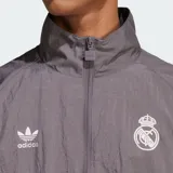 Chaqueta deportiva Adidas del Real Madrid en color gris, con diseño de bloques en blanco y azul marino, cuello alto, cierre frontal y las icónicas tres tiras en las mangas.