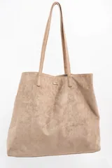 Bolso tipo tote de gran tamaño confeccionado en gamuza sintética color tostado. Presenta un diseño minimalista con asas largas de hombro y una pequeña placa metálica frontal con el logo de la marca.