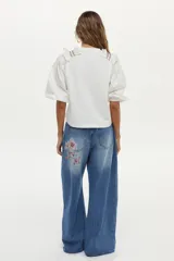 Blusa blanca con cuerpo de jersey y mangas de tela plana abullonadas. Cuenta con volados en los hombros decorados con bordados en contraste.