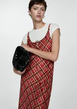 Vestido largo de tirantes finos con estampado de cuadros en tonos rojizos y marrones. Presenta un diseño recto, tejido fluido y un lazo decorativo en el escote.
