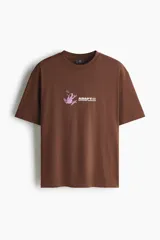 Remera de manga corta color marrón, con cuello redondo y corte holgado. Presenta un pequeño estampado en el pecho con la figura de una rana en color lila y la inscripción "ADAPTO OUTDOOR SUPPLIES".