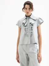 Camisa de cuero ovino color gris claro, con cuello camisero, volados centrales y mangas cortas con volumen generado por frunces en la copa. Cierre frontal con broches forrados.