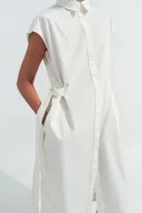 Vestido midi camisero de algodón color negro, con cuello solapa, sin mangas y cierre frontal con botones ocultos por solapa. Incluye cintura ajustable con tiras de lazada lateral en el mismo tejido.