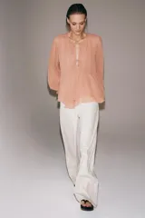 Blusa de viscosa color rosado con cuello redondo, manga larga abullonada con puños elásticos y cierre frontal con lazadas. Presenta un diseño con volantes en el bajo y corte asimétrico.