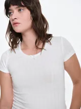 Remera de manga corta entallada en tejido pointelle, con escote redondo y detalle de picot con lazo pequeño en el centro. Diseño ajustado al cuerpo con textura acanalada.
