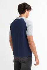 Remera tipo polo de manga corta con diseño combinado, cuerpo en color gris y mangas y cuello en color azul marino. Presenta cierre frontal con botones y detalles de líneas finas en el cuello.