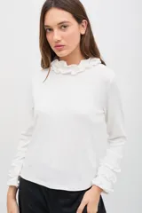 Polera blanca de manga larga con cuello alto y puños decorados con volados. Confeccionada en tejido suave con textura sutil.