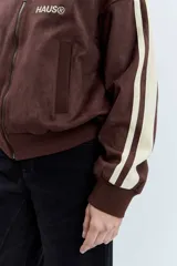 Cazadora de estilo deportivo en color marrón, confeccionada en tejido tipo gamuza. Presenta cierre frontal completo, cuello alto, puños y ruedo acanalados, y franjas laterales en tono rosado. Incluye el logo de la marca bordado en el pecho.