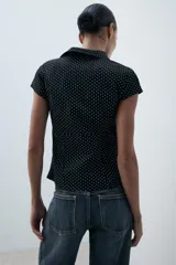 Camisa entallada de popelín negro con estampado de lunares blancos, escote en pico, cuello solapa y mangas muy cortas. Cuenta con costuras marcadas y cierre lateral con cremallera oculta.