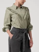 Blusa de algodón orgánico color verde oliva, con cuello camisero, cierre frontal de botones y mangas largas. Presenta detalles de volantes bordados en el frente y bordados calados en toda la prenda.