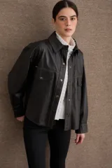 Camisa de cuero negro con costuras a la vista en color contrastante, cierre frontal con botones metálicos a presión, bolsillos frontales con solapa y mangas largas con puños abotonados.