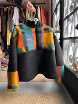 Buzo canguro de corderito con cierre frontal parcial y cuello alto. Diseño en bloques de color con estampado a cuadros en tonos verde, amarillo, rojo y azul en la parte superior y mangas, y tejido negro texturizado en la parte inferior.