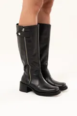 Bota estilo biker de cuero negro con caña alta y cierre metálico lateral. Presenta un diseño versátil con estribo y cadena desmontables, taco cuadrado y suela con plataforma.