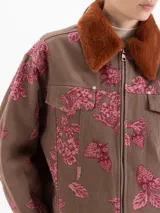 Chaqueta de gabardina color marrón con estampado floral en tonos rosados, corte recto y cierre frontal con cremallera. Presenta cuello de piel sintética en color tostado y bolsillos frontales con solapa.