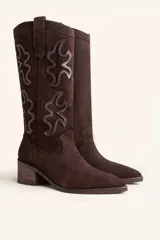 Bota de caña alta estilo tejano confeccionada en gamuza color marrón oscuro. Presenta detalles de costuras decorativas en forma de llama en la caña, taco cuadrado de madera y punta almendrada.