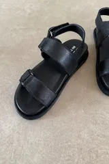Sandalia plana color marrón de cuero vacuno, con tres tiras ajustables con hebillas y cierre de velcro, y base acolchada.