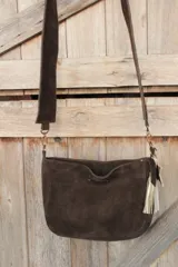 Bandolera confeccionada en cuero de gamuza marrón, con cierre superior. Presenta un llavero colgante con forma de estrella revestido en tachas metálicas y tiras flecadas en tono dorado.