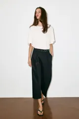 Pantalón de popelina de tiro alto y pernera ancha, de color negro. Presenta pinzas delanteras, bolsillos laterales y falsos bolsillos de vivo en la espalda. Incluye un cinturón combinado tipo pañuelo trenzado con tiras colgantes y cuentas de madera. Cierre frontal con cremallera, botón interior y gancho.