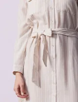 Vestido camisero corto a rayas verticales celestes y blancas, con cuello con solapa, abertura frontal con botones, dos bolsillos de parche en el pecho y lazo de ajuste en la cintura.