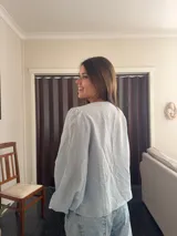 Camisa de tela liviana color celeste, con escote en V, mangas largas abullonadas con puños elásticos y detalle de volado fruncido en el frente con cierre de cordones ajustables.