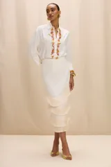 Camisa de color blanco hueso con cuello clásico, mangas tres cuartos y corte recto. Presenta un aplique decorativo frontal compuesto por piedras de colores y detalles metálicos dorados.