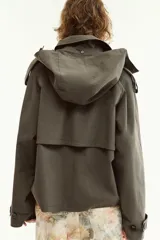 Trench corta verde militar con capucha, cuello solapa y manga larga con trabillas en los puños. Presenta bolsillos delanteros de vivo y cierre frontal cruzado con doble botonadura.