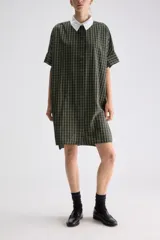 Vestido camisero midi de calce relajado a cuadros en tonos verde y negro, confeccionado en algodón liviano. Presenta cuello clásico en contraste de color blanco, cartera frontal oculta con botones y mangas cortas con vuelta.