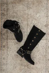 Botas altas de gamuza negra con caña alta, detalle de tachas metálicas laterales y correa con hebilla en el tobillo.