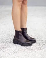 Bota tipo Chelsea de cuero color marrón oscuro, con paneles elásticos laterales en color negro y tirador trasero. Posee una suela de goma tipo track con relieve y costura blanca decorativa en el cerco. Hecha a mano en Uruguay.