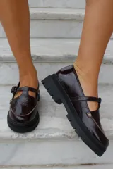 Zapato tipo guillermina en cuero charol color bordó, con suela track negra de goma, cierre con hebilla ajustable y detalle de herrajes metálicos dorados.