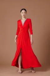 Vestido largo rojo de corte elegante con escote en V profundo adornado con pedrería brillante. Presenta mangas tres cuartos con puños decorados con pedrería, detalle de nudo frontal en la cintura y una abertura lateral en la falda que aporta movimiento.