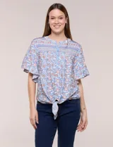 Blusa holgada de poliéster con estampado floral y geométrico en tonos celestes, azules y anaranjados sobre base blanca. Presenta cuello a la base con escote en V y botones frontales, mangas cortas amplias de estilo mariposa y un nudo en el ruedo.