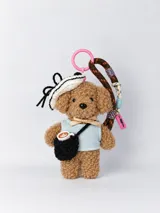 Llavero colgante con diseño de ovejita de peluche marrón vestida con chaleco celeste y moño dorado, sombrero de punto blanco y negro con lazo, y una mini cartera negra con una taza de café en miniatura. Incluye correa de cordón marrón estampado y anillo plástico rosa.