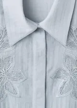 Blusa de tejido ligero color celeste con cuello camisero y cierre frontal de botones ocultos. Presenta mangas largas con puños anchos y detalles florales bordados en la parte superior delantera.