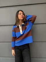 Buzo tipo rugby de algodón con diseño de rayas horizontales anchas en azul y marrón. Presenta cuello polo en color crema con cierre frontal de tres botones y mangas largas.