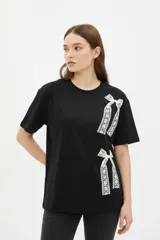 Remera negra de algodón con cuello redondo, manga corta y corte holgado, decorada con un estampado de moños de encaje blanco en el frente.