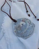 Collar artesanal con dije central de metal con textura orgánica irregular que evoca una ostra. El dije está montado sobre un cordón de algodón natural, permitiendo un uso versátil y regulable.