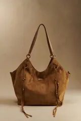 Bolso tipo shopper de gamuza vegana color marrón, con tiras colgantes decorativas a los lados y placa metálica frontal con el logo de la marca. Cuenta con asas largas de hombro.